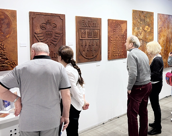 Besucher der Kunstmesse artfair in Köln betrachten rostfarbene Reliefbilder und moderne Metallkunst an einer weißen Galeriewand. Die ausgestellten Werke zeigen abstrakte Strukturen, Wappen und texturierte Oberflächen im Industrial- und Rost-Design.