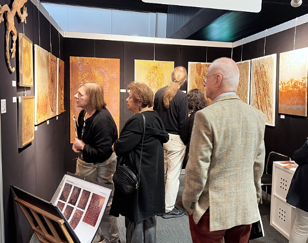 Mehrere Menschen stehen auf einer Kunstmesse in Masstricht vor abstrakten Kunstwerken mit warm beleuchteten Gemälden in Braun-, Gold- und Rosttönen an schwarzen Wänden. 